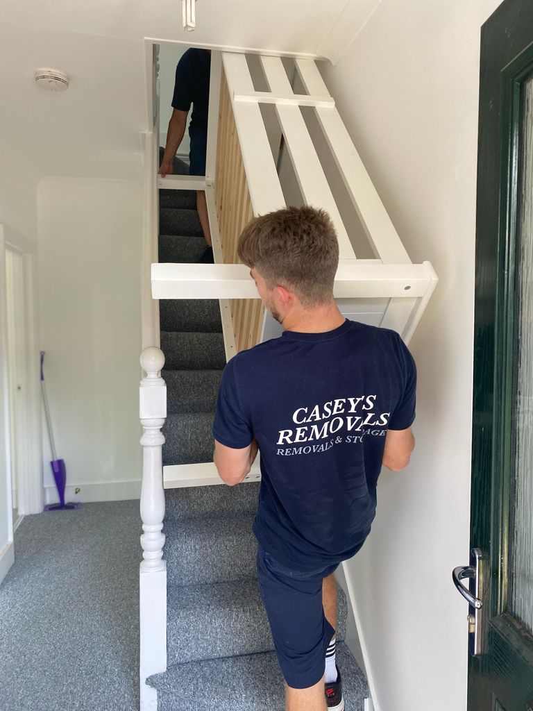 Removal men on stairs carrying bed frame wearing blue t-shirt with Casey's Removals printed on it