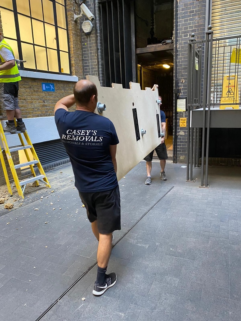 Men carrying desk from office building wearing a dark tshirt with casey's removals printed on back