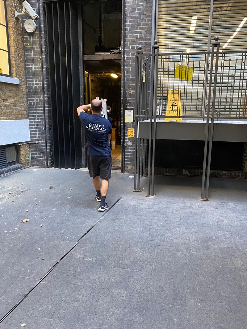 Men carrying office furniture outside of office building wearing a dark tshirt with casey's removals printed on back